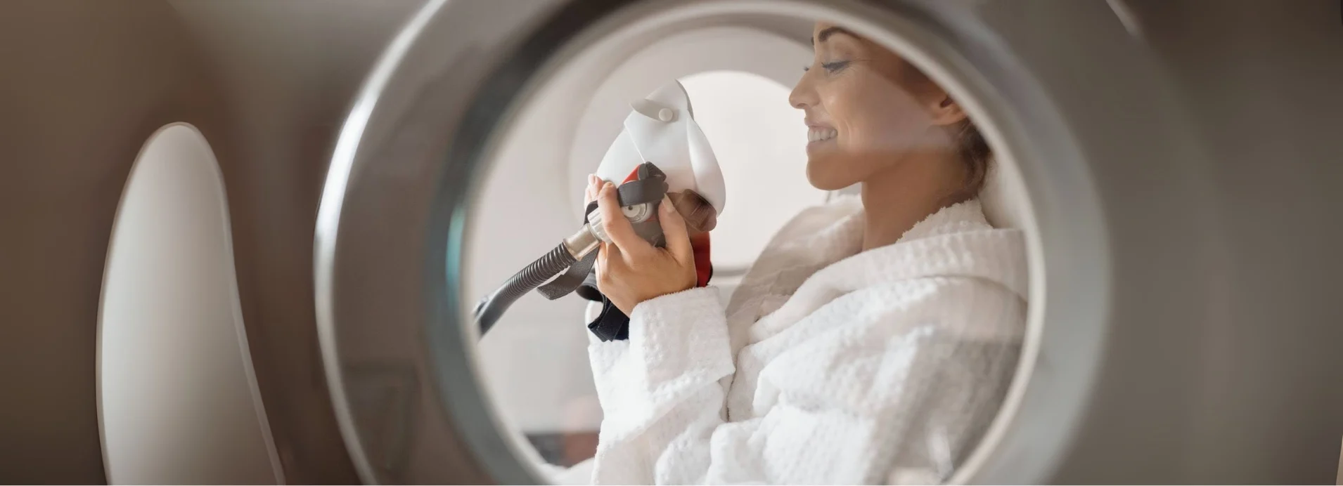 Woman wearing a white robe smiles as she applied a breathing mask inside a hyperbaric oxygen therapy chamber