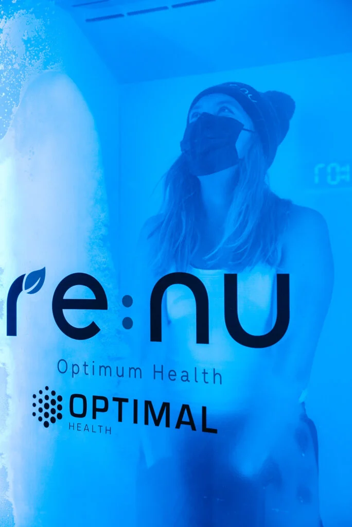 Woman wearing a woolly hat and face covering standing in the Re:nu Optimum Health cryotherapy chamber looking up.