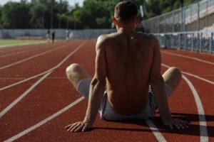 Male athlete seen from behind sitting on a running track, stretching his legs after training in London
