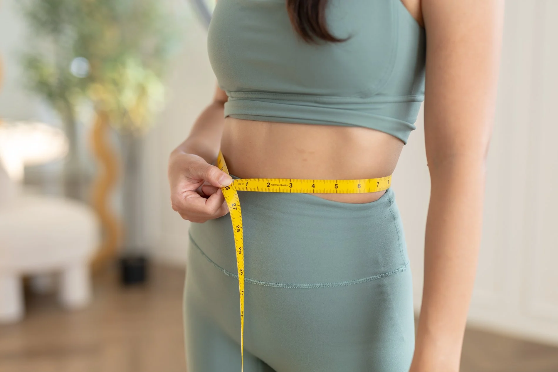 A woman in fitness wear tracking weight loss treatment progress by measuring her waist with a tape measure.